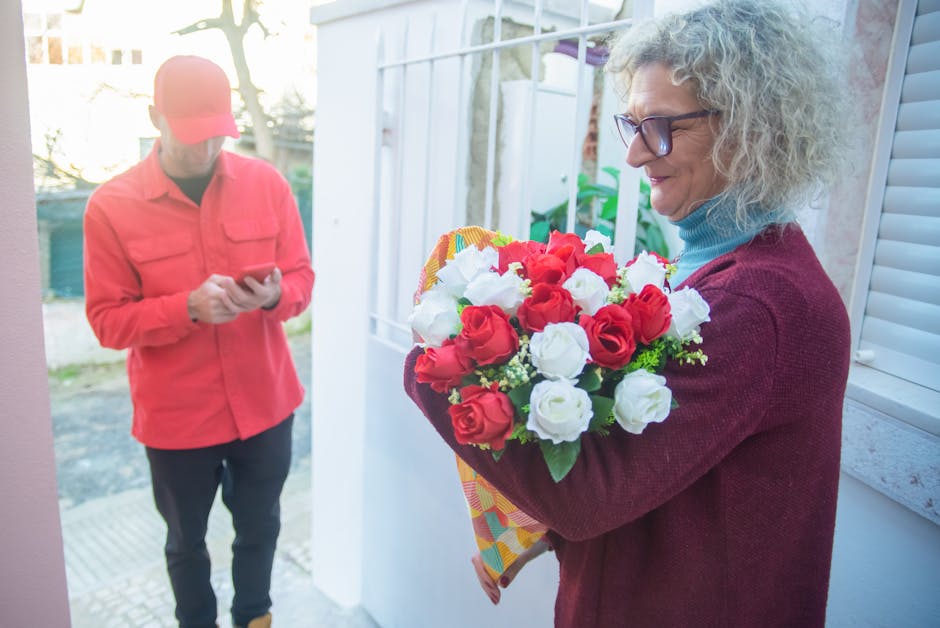 A woman with curly gray hair and glasses is holding a bouquet of fresh red and white roses wrapped in colorful patterned paper, standing near a bright window. A person in a coral jacket and black pant