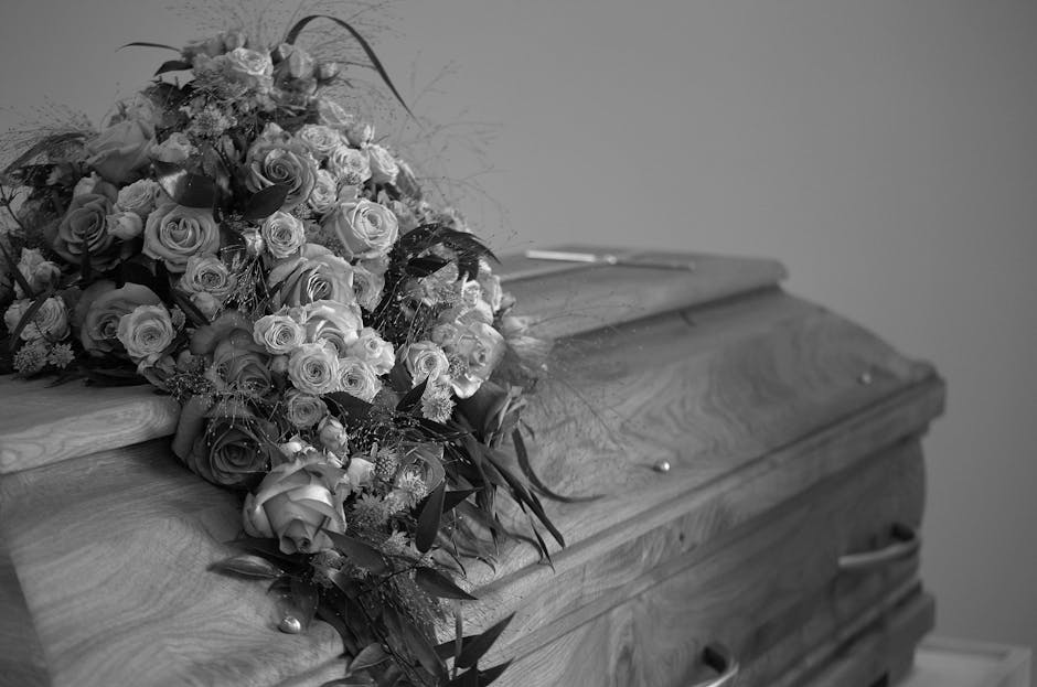 A large bouquet of fresh flowers resting on a wooden coffin, featuring roses and other blooms in various stages of openness, with some petals showing intricate textures. The flowers include light-colo