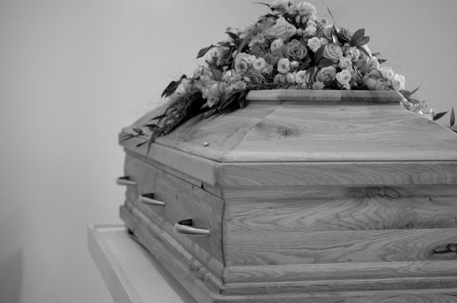 A wooden coffin with a polished finish rests on a stand, adorned with a large floral arrangement on top. The bouquet features a mixture of roses, carnations, and other flowers in soft pastel shades, a