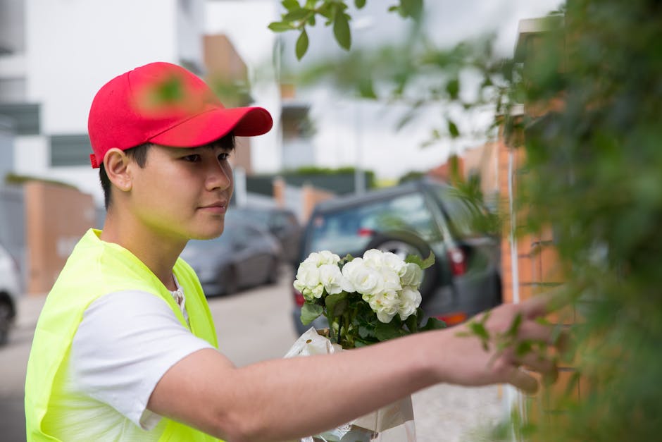 A young delivery person wearing a red cap and a high-visibility yellow vest is placing a bouquet of fresh white hydrangeas wrapped in light paper onto a doorstep. The arrangement features large, lush 