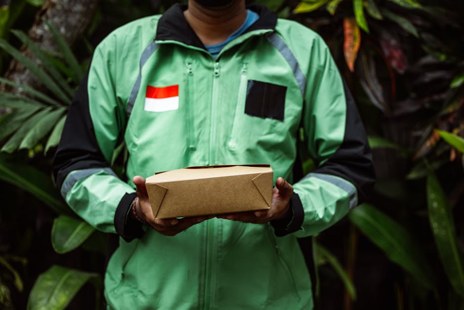 A delivery person wearing a green and black jacket with a red and white flag patch and black accents is holding a rectangular brown cardboard box with folded edges. The individual is standing outdoors