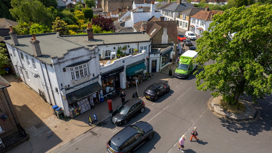 An aerial view of a small commercial street in Stamford Hill, N16, showing a white building housing a flower shop with a teal awning. The shop displays floral arrangements outside, and a delivery van