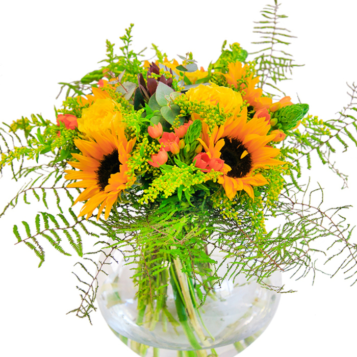 The first impression of this bouquet is one of jubilant abundance: sunflowers in radiant yellow, their chocolate-brown centers capturing light with every angle, are nestled close to golden garden roses whose petals unfurl in plush spirals. The arrangement mirrors the vibrant life of Stamford Hill's high streets and green spaces, as deep emerald leaves and trailing feathery ferns spill over the edges of a pristine, round glass vase. Among the bold blooms, shiny Hypericum berries-bright red as summer cherries-sparkle with cheerful intent, their glossy skins contrasting with the matte finish of the rose and sunflower petals. Threaded throughout are subtle sprigs of pale, yellow-tipped filler, likely goldenrod or solidago, and slender green flower spikes that add both height and softness to the design, evoking the carefree beauty of community gardens in the area. Bluish eucalyptus leaves peek through, suggesting a modern, rustic touch beloved by Stamford Hill's florists. Up close, the layers of texture come alive: the sunflowers' suede-like petals, the smooth rose buds, the crisp edges of fresh greenery, and the delicate touch of ferns that flutter at the slightest breeze. The clear glass vase cradles every luminous stem, its transparency highlighting the arrangement's carefully tuned architecture and enhancing its sense of clean, contemporary brightness. Subtle natural scents-soft rose, sweet earth, and the faintest herbal note-mix together, recalling the fresh air of Alexandra Palace parklands nearby. This bouquet is perfect for marking special moments-a birthday surprise, an anniversary dinner, or any celebration where happiness takes centre stage in a Stamford Hill home.
