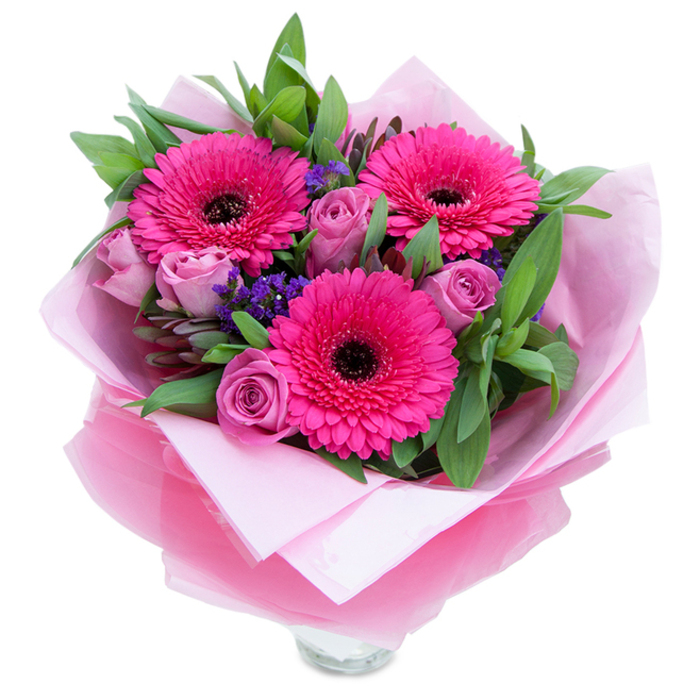 I picture three vivid fuchsia Gerbera daisies as the confident visual anchors in this bouquet, their layered petals radiating outward and framing deep, near-black centres that give the arrangement a beautifully grounded focal point. Around them, dusty-lavender roses open in measured stages-petals curled and soft, emitting a gentle, classic rose perfume that mingles with the faint green sweetness of the gerberas. Interspersed clusters of purple statice add a slightly papery texture and a cool violet edge that complements the pinks without overpowering them. Underpinning the blooms is a generous sweep of green foliage-broad, slightly waxy leaves resembling salal or eucalyptus-that offers both structural support and a fresh, forest-floor aroma. The bouquet is carefully cocooned in an inner sheet of pale pink matte tissue and an outer veil of translucent fuchsia-pink cellophane, the latter catching light with a subdued shine; the wrapping is folded with thoughtful precision, indicating skilled hands. The lower rim of a clear glass vase is visible, suggesting the stems are arranged for immediate display on a dining table or hallway console. The photograph captures soft, even lighting that reveals petal texture-the sheen of the gerbera petals and the satiny depth of the roses-without stark shadows. As a florist familiar with Stamford Hill and neighbouring Stoke Newington, I see this composition as perfectly suited for intimate celebrations: birthdays, Mother's Day, anniversaries, or a tender 'thinking of you' gift that carries both cheer and quiet elegance.