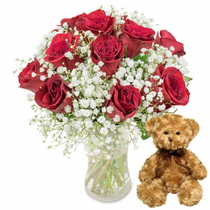 This image portrays a tender, romantic gift set arranged as if ready for a same-day delivery across Stamford Hill and the leafy avenues leading toward Stoke Newington. Dominating the left side is an elegant bouquet of around a dozen deep crimson red roses, neatly gathered in a clear cylindrical glass vase. The roses are lush and full, their velvety petals layered in tight spirals that gradually open into broad, cup-shaped blooms. Each flower varies slightly in its stage of openness, which adds natural rhythm and depth to the arrangement. Encircling and weaving between the roses is a generous spray of white baby's breath (Gypsophila), its countless tiny blooms forming soft, airy clouds that contrast beautifully with the intense red. The crisp white and deep green foliage frame the roses without distracting from their bold, romantic statement. Through the glass vase, you can see the cleanly cut green stems submerged in clear water, suggesting the flowers have been freshly prepared. To the right of the vase sits a charming teddy bear, about the height of the vase, with light brown plush fur that looks invitingly soft to the touch. The bear sits upright, paws resting loosely in front, giving it a relaxed and approachable pose. Its small black button eyes, dark nose, and gentle stitched smile create a sweet, affectionate expression, while a narrow dark brown satin ribbon is tied in a simple bow around its neck. With its uncluttered white background and balanced composition, the scene evokes the feeling of a carefully chosen anniversary or Valentine's gift, the kind of thoughtful surprise a local customer might order from Flower Delivery Stamford Hill for a loved one living near Clapton Common or along Amhurst Park.