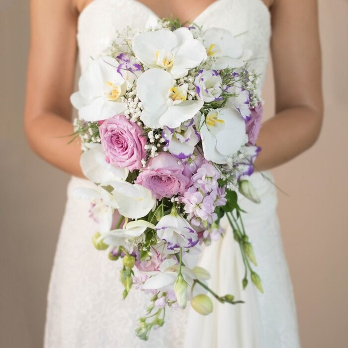 In this atmospheric photograph taken near the leafy avenues of Stamford Hill, a bride stands against a softly blurred backdrop in shades of cream and beige, hands poised in gentle embrace around an extravagant cascading wedding bouquet. The artistry of the arrangement is evident in its teardrop silhouette, overflowing with large, immaculate white phalaenopsis orchids-their broad petals unfurl with a silky sheen, accentuated by subtle yellow-and-violet centers. Among the orchids nestle romantic, blush-tinted garden roses, their tightly packed, plush petals suggestive of English summer gardens and the timeless beauty cherished by many couples in Stamford Hill. Subtly spiky lavender stock provides texture and a whisper of pastel purple, while playful, ruffled lisianthus blooms reveal delicate white petals edged in deep lilac, echoing the vibrant flower markets often visited by locals along Amhurst Park. Wispy sprigs of baby's breath weave throughout, contributing an airy lightness, while understated green foliage creates a natural, hand-gathered feel. The composition draws the senses towards the bride's elegantly detailed lace gown, whose bodice and skirt appear through the lower frame, their lacework echoing the intricate craftsmanship of the bouquet. Light and petals intertwine in harmonious balance, crafting a sense of serene sophistication and romance perfect for a Stamford Hill wedding. This bouquet is not merely a floral accessory-it is an evocative centerpiece, brought to life by a florist intimately familiar with the tastes and traditions of this diverse neighbourhood.