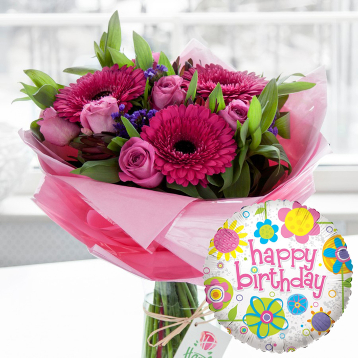 A joyful birthday scene unfolds in this image, where a luxurious pink floral arrangement and a festive balloon sit together as if waiting to be carried to a party in Stamford Hill. Dominating the composition is a clear cylindrical vase generously filled with a hand-crafted bouquet. Large Gerbera daisies in rich fuchsia and magenta take centre stage, their petals forming perfect circles around deep, almost black centres that add striking contrast. Around them, layers of delicate pink roses in varying stages of bloom - from tight buds to half-open spirals - introduce softness and a gentle romantic note. Threaded among these feature flowers are clusters of small, deep purple filler blooms, likely statice, which add a subtle, cloud-like texture and intensify the colour palette. A lush frame of vibrant green foliage cradles the bouquet, the leaves glossy and full, giving the whole arrangement a sense of freshness, as though it has just been prepared by a local florist for a home near Stamford Hill station or the leafy streets off Cazenove Road. The stems are wrapped at the base in layers of translucent light pink cellophane that fan upward, creating a gossamer-like halo that catches and diffuses the light. A rustic length of twine is tied in a simple bow around the neck of the vase, holding a small off-white tag printed with an elegant rose motif, a quiet nod to quality and attention to detail. In the lower right foreground, a round Mylar Happy Birthday balloon adds a playful flourish. Its crisp white surface, lightly flecked with grey, is scattered with lively floral and circular designs in bright yellow, sky blue, lime green, vivid pink and deep purple. Across the centre, happy birthday is written in bold pink script, echoing the colours of the flowers. The softly blurred indoor background, illuminated by natural light streaming from a nearby window, keeps the focus firmly on this thoughtful, ready-to-deliver birthday gift pairing.