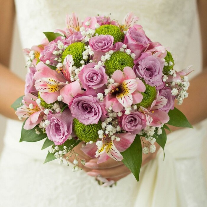 An exquisite bridal bouquet, brimming with delicate detail and soft colour, is lovingly held against the lace of a flowing white dress-its artistry a tribute to Stamford Hill's floral heritage. Meticulously arranged, this spherical bouquet features velvety roses in shades of powder pink and gentle lavender, their petals fresh and adorned with glistening droplets, reminiscent of a spring morning on Amhurst Park. Vibrant lime green button chrysanthemums introduce a lively, modern edge, contrasting beautifully with the pastel hues and adding a spirit of renewal to the design. Interspersed are graceful pink alstroemeria, their trumpet-shaped petals streaked with maroon and gold, evoking the rich tapestry of cultures found in Stamford Hill's bustling markets. Ethereal clusters of white baby's breath float like soft clouds among the larger blossoms, contributing volume and a sense of lightness. Deep green rose leaves emerge at the base, framing the bouquet with structure and depth. The bride's hands, adorned with a shimmering wedding band, cradle the bouquet gently, as though holding a promise of happiness yet to unfold. Crafted by a Stamford Hill florist with a reputation for excellence, this bouquet is perfect for couples looking to marry classic romance with a hint of contemporary flair, ideal for spring or summer celebrations in this vibrant London community.