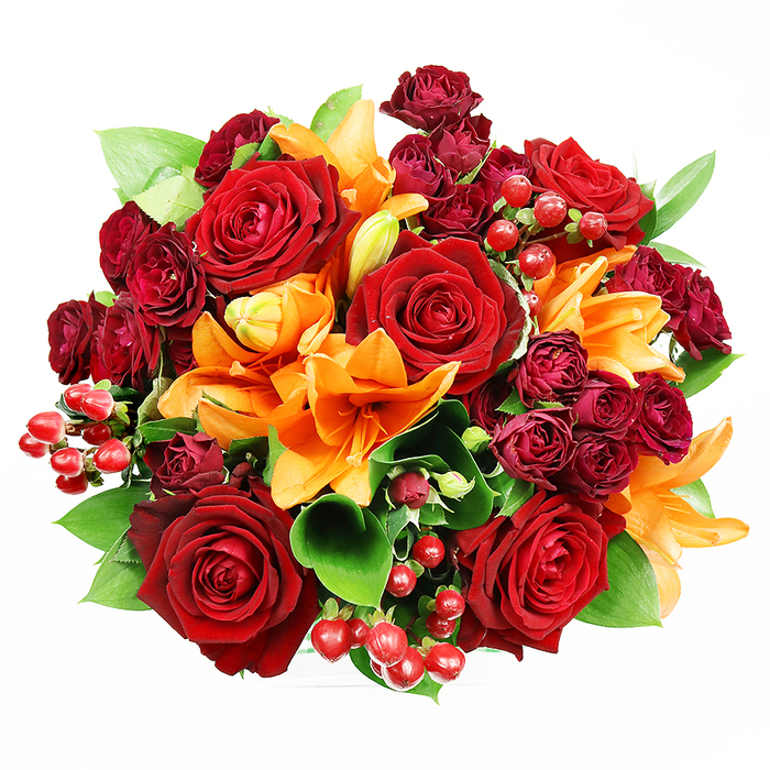 Captured in an overhead view on a spotless white surface, this opulent, circular bouquet reads like a miniature, cultivated garden of warm tones and rich textures. Dominating the arrangement are several large, fully blown deep red roses whose velvety petals spiral outward, imparting classic romance and a tactile sense of depth. Interspersed among these are brilliant orange Asiatic lilies, their broad trumpet petals sometimes open to reveal dark freckles and prominent stamens, sometimes tucked into graceful buds - a contrast that suggests both immediacy and unfolding promise. Tight clusters of deep burgundy spray roses add jewel-like density and a velvet finish that deepens the overall colour story, while neat sprays of glossy red hypericum berries glint like tiny rubies, introducing lively points of light and playful texture. Lush green foliage, likely ruscus or salal, peeks through to ground the palette with fresh, cooling tones. The lighting is gentle and even, lifting petal surfaces and berry sheen without casting hard shadows; the arrangement feels both luxurious and welcoming. This is the kind of bouquet Flower Delivery Stamford Hill would send for milestone anniversaries, warm seasonal celebrations, or as a richly toned home accent for a flat near Stamford Hill Road or a parlor overlooking Clapton Common. The scent you imagine is a soft chorus of rose depth with a bright, spicy note from the lilies - a composition that feels handcrafted, balanced, and ready to make an impression.