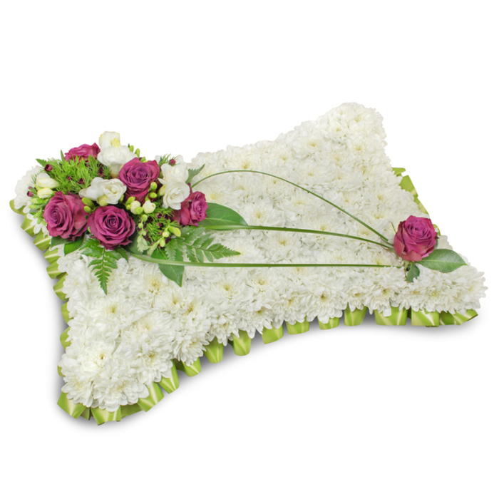 A classic funeral pillow arranged with meticulous care, presented on a white background that highlights texture, colour and form. The main surface is densely padded with small, immaculate white chrysanthemum heads, their countless petals interlocking to create a soft, cloud-like cushion suggestive of rest and serenity. Along the lower rim a neatly folded lime-green satin ribbon wraps the pillow, the subtle shimmer of the fabric forming a pleated frame that contrasts with the chrysanthemums' velvety matt finish. Anchoring the composition in the top-left corner is a vibrant cluster of deep fuchsia roses; each rose opens with luxuriant, velvety layers, set amid delicate white freesia and creamy lisianthus blooms that introduce an airy fragrance and delicate texture. Lush green foliage - including feathery fern fronds and glossy leaves - fans out behind the cluster, giving a natural backdrop and weight to the focal blooms. Elegantly arcing across the floral surface are three slim blades of dark green bear grass, their graceful curves leading the eye from the main cluster toward the opposing corner where a solitary fuchsia rose sits thoughtfully among leaves, echoing the palette and completing the visual rhythm. The composition casts a soft shadow on the white surface beneath, enhancing the sense of depth and the pillow's sculptural quality. Favoured for memorial services and chapel tributes across Stamford Hill and nearby neighbourhoods, this arrangement reads as both a comforting emblem of sleep and a dignified token of remembrance, made with the attentive craftsmanship of an experienced florist.