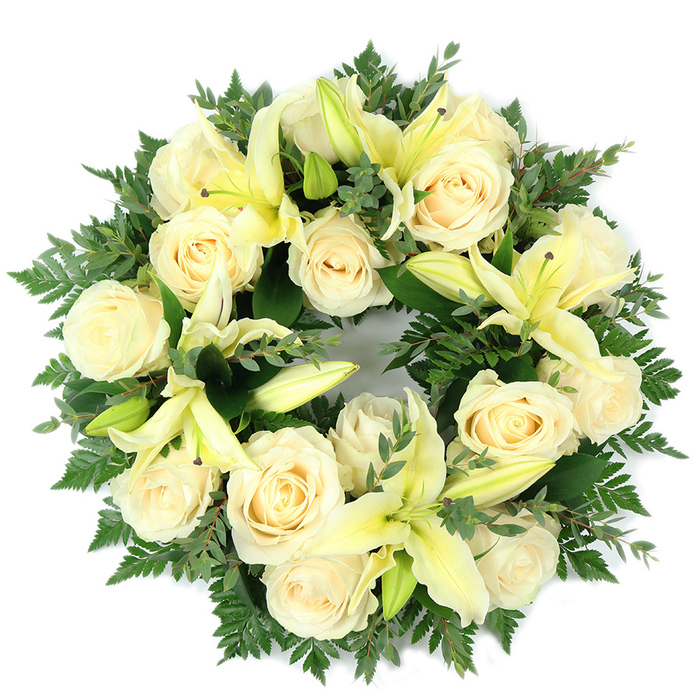 A meticulously arranged circular wreath laid flat and photographed from above, composed of creamy white roses and pale lemon lilies set against a lush bed of green foliage. The wreath's density is its strength: close-packed ivory roses-fifteen or so at the core-display classic, multi-layered petals that glow softly under neutral light, while the pale yellow trumpet lilies punctuate the composition with graceful arcs; some lilies are fully open, others poised as buds to introduce visual cadence and the sense of future bloom. The greens form a rich, supportive base-delicate fern fronds lending feathery edges and broader, rounded leaves similar to eucalyptus providing depth and contrast-so that the light-coloured blooms float above a verdant stage. The circle's symmetry and the choice of gentle tones create a feeling of calm, a visual language of peace and purity appropriate for remembrance. Sensory cues remain vivid in description: the subtly sweet, powdery perfume of the roses layered with the fresher, green-tinged scent of lilies, plus the cool, tactile feeling of the foliage beneath. Presented against a clean white backdrop, the wreath reads as an elegant memorial piece, suitable for a funeral service, graveside tribute, or as a heartfelt expression of condolence; it is the sort of thoughtful arrangement you might commission from a trusted local florist for a service in Stamford Hill or a nearby church or chapel.